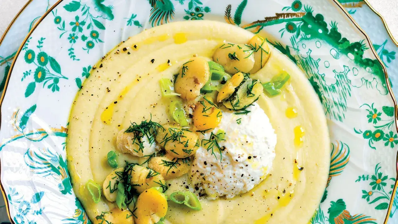Un cremoso macco di fave fresche, guarnito con ricotta, fave intere, cipollotto e aneto, servito su un piatto decorato.