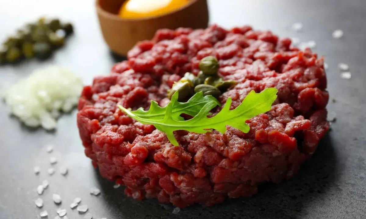 Tartare di manzo fresca, guarnita con capperi e rucola, pronta per essere gustata con tuorlo d'uovo e cipolla.