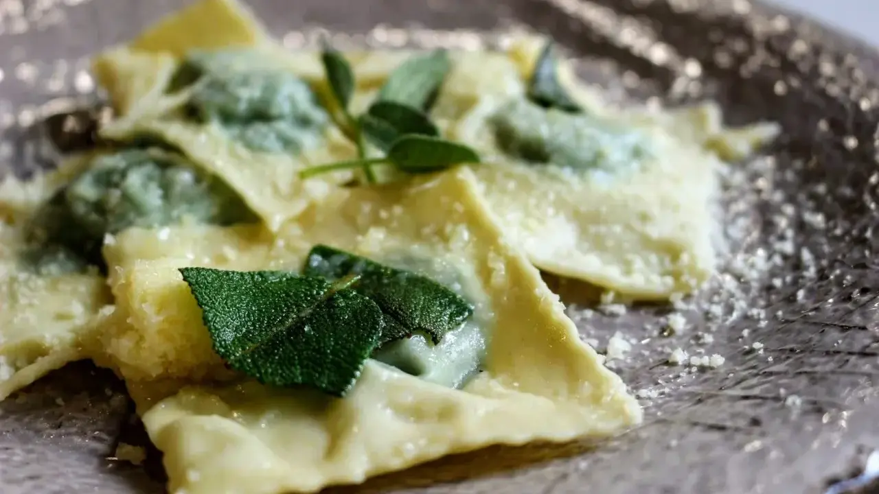 Ravioli ricotta e spinaci, conditi con burro fuso e foglie di salvia fresca, spolverati di parmigiano grattugiato su un piatto elegante.