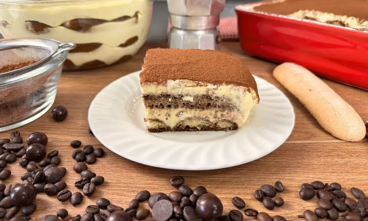 Una fetta di tiramisù con uova pastorizzate, cosparso di cacao, su un piatto bianco, circondato da chicchi di caffè e cioccolatini.