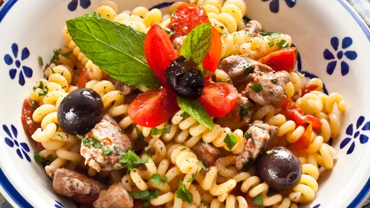 Pasta con pesce spada, pomodorini, olive nere e menta fresca in una ciotola decorata.