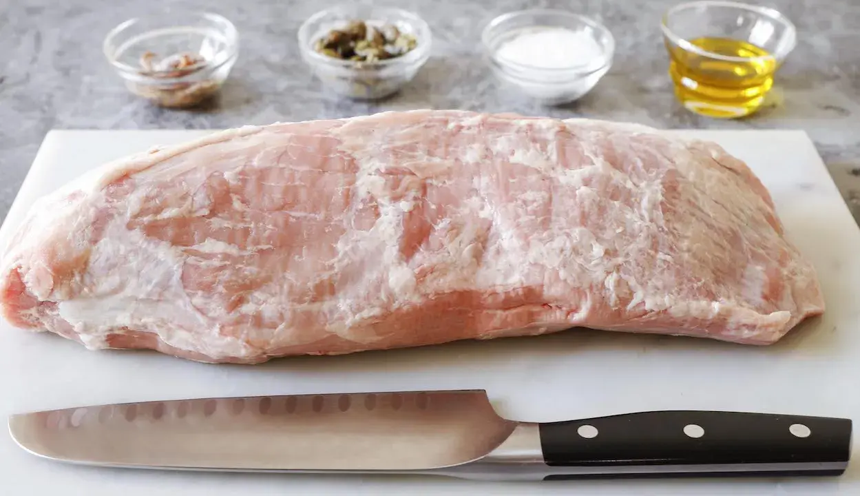 Un pezzo di girello di vitello crudo, pronto per essere cucinato, con coltello e ciotoline di condimenti sullo sfondo.