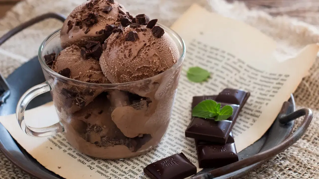 Coppa di gelato al cioccolato con scaglie di cioccolato fondente e foglioline di menta, su un vassoio arrugginito con un libro aperto.