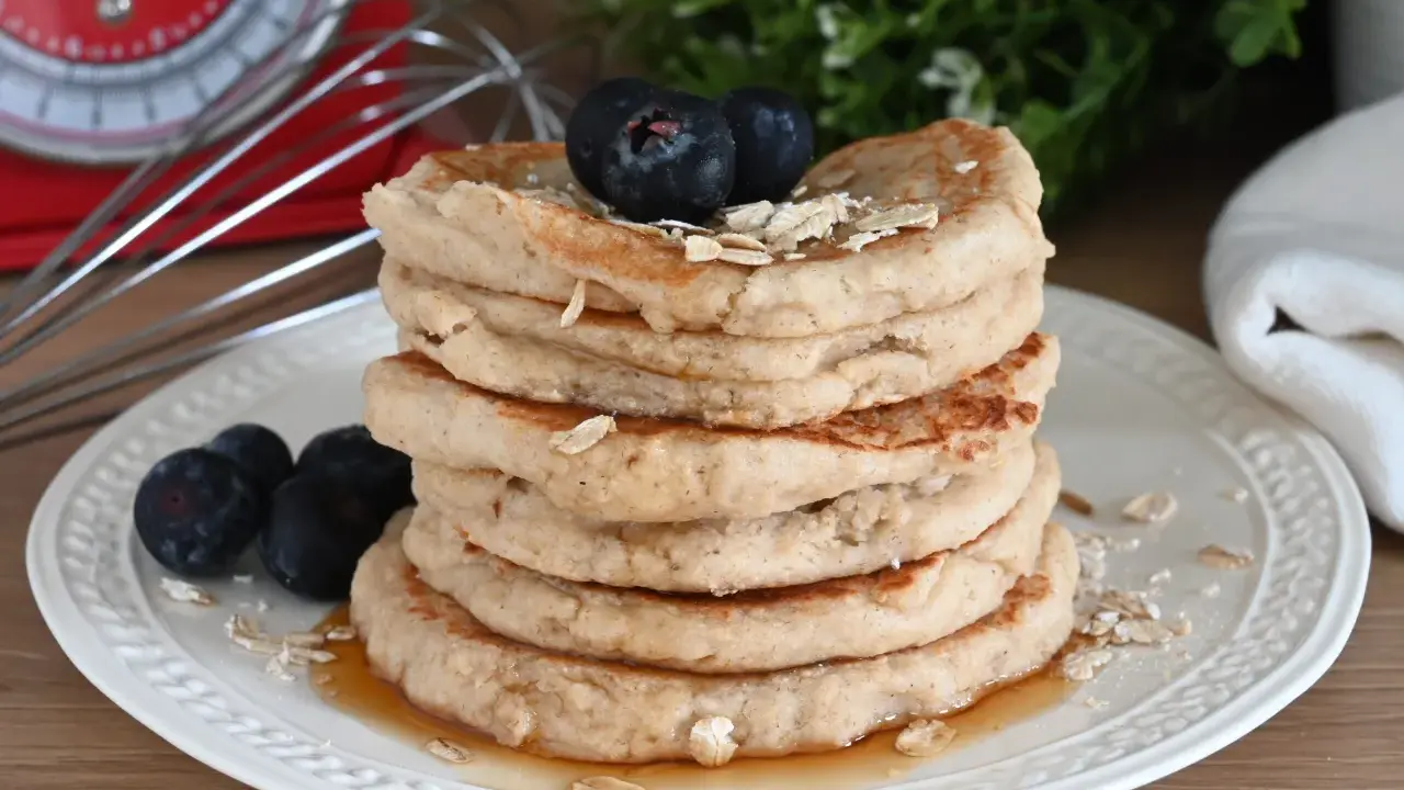 Pila di pancake con farina di avena, guarniti con mirtilli e fiocchi d'avena, su un piatto bianco con sciroppo d'acero.