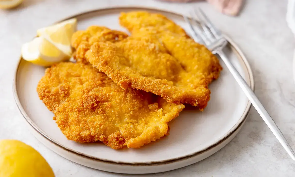 Due cotolette di pollo dorate e croccanti, servite su un piatto con spicchi di limone e una forchetta.