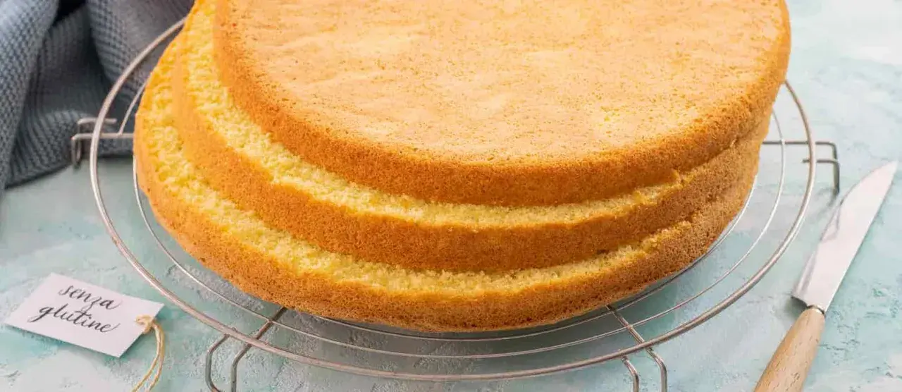 Tre soffici strati di pan di spagna senza glutine raffreddano su una griglia. Un'etichetta con scritto "senza glutine" è appesa alla griglia.