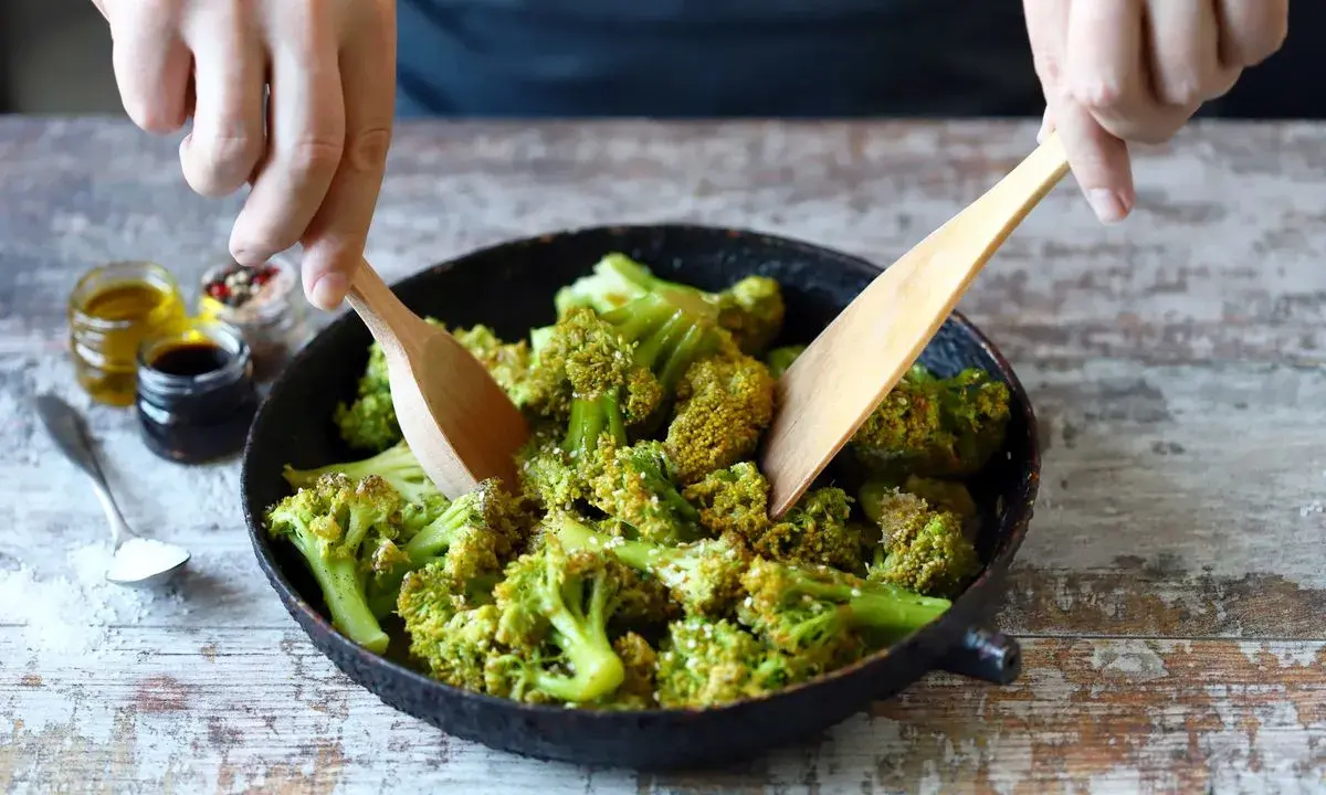 Mani che mescolano broccoli in padella con due palette di legno. Sullo sfondo, piccoli barattoli con olio, aceto balsamico e spezie.