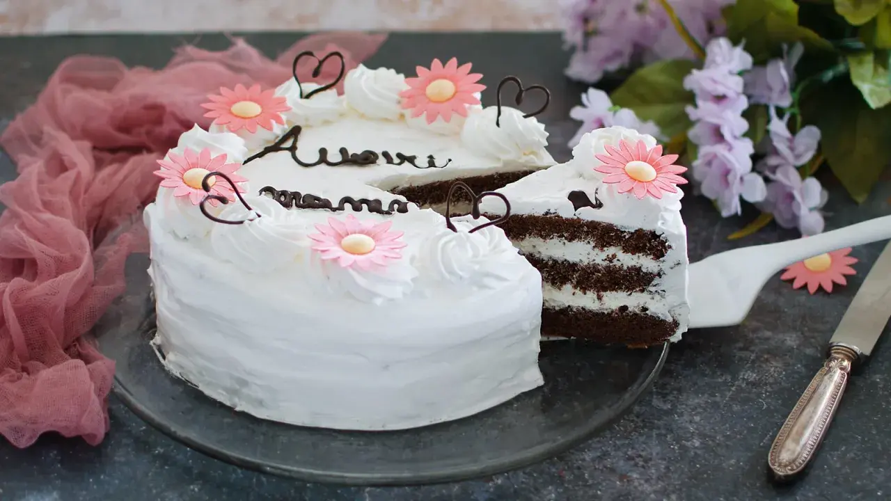 Una torta festa della mamma con glassa bianca, fiori rosa e cuori di cioccolato. La fetta tagliata rivela strati di torta al cioccolato e crema.