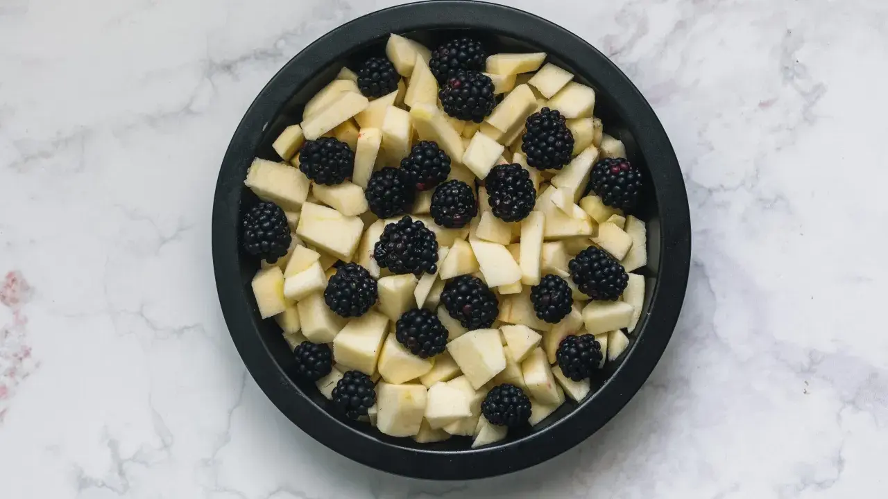 Mele a cubetti e more in una teglia nera, pronte per un delizioso crumble di mele.