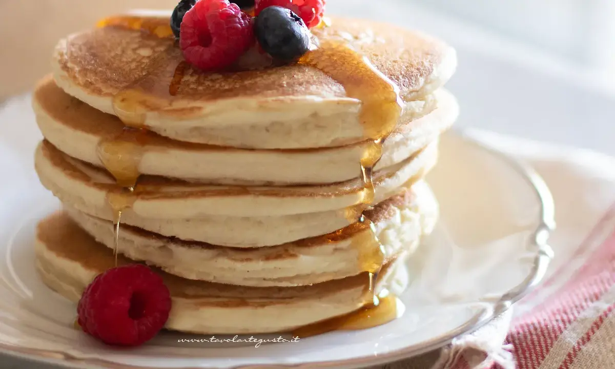 Pancakes senza glutine dorati, impilati e inzuppati di sciroppo, guarniti con lamponi e mirtilli freschi.