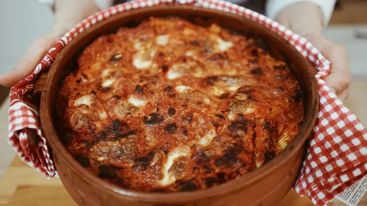 Un invitante pasta al forno appena sfornata, con crosta dorata e ripieno filante, tenuta con un canovaccio a quadretti rossi.