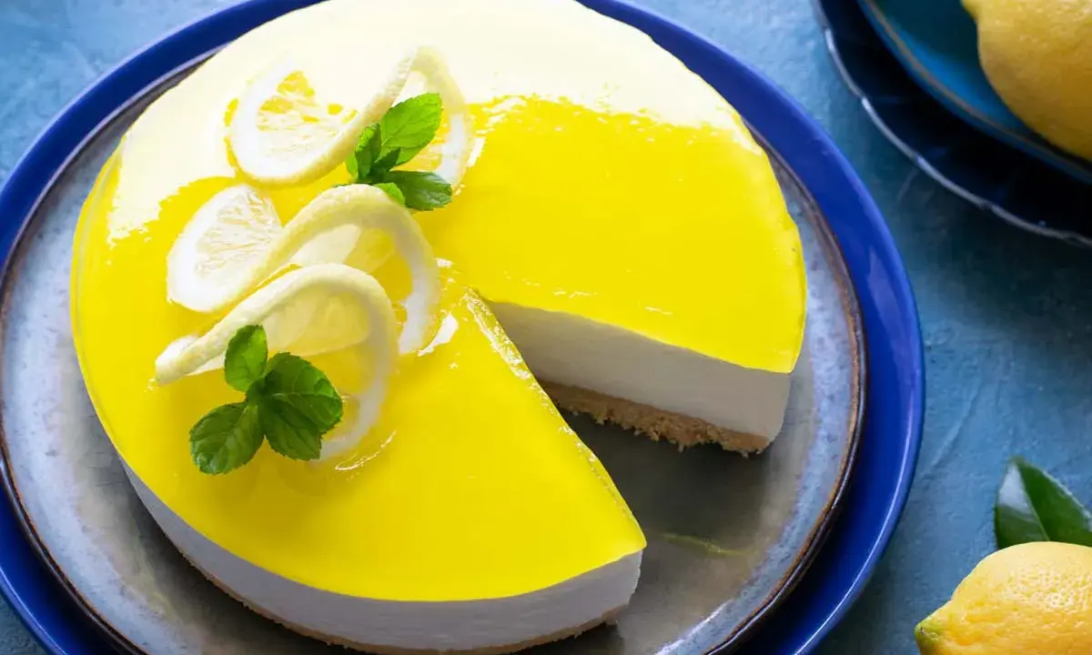 Delizioso cheesecake al limone con glassa gialla brillante, decorato con fette di limone e menta. Una fetta è stata tolta, rivelando la base croccante.