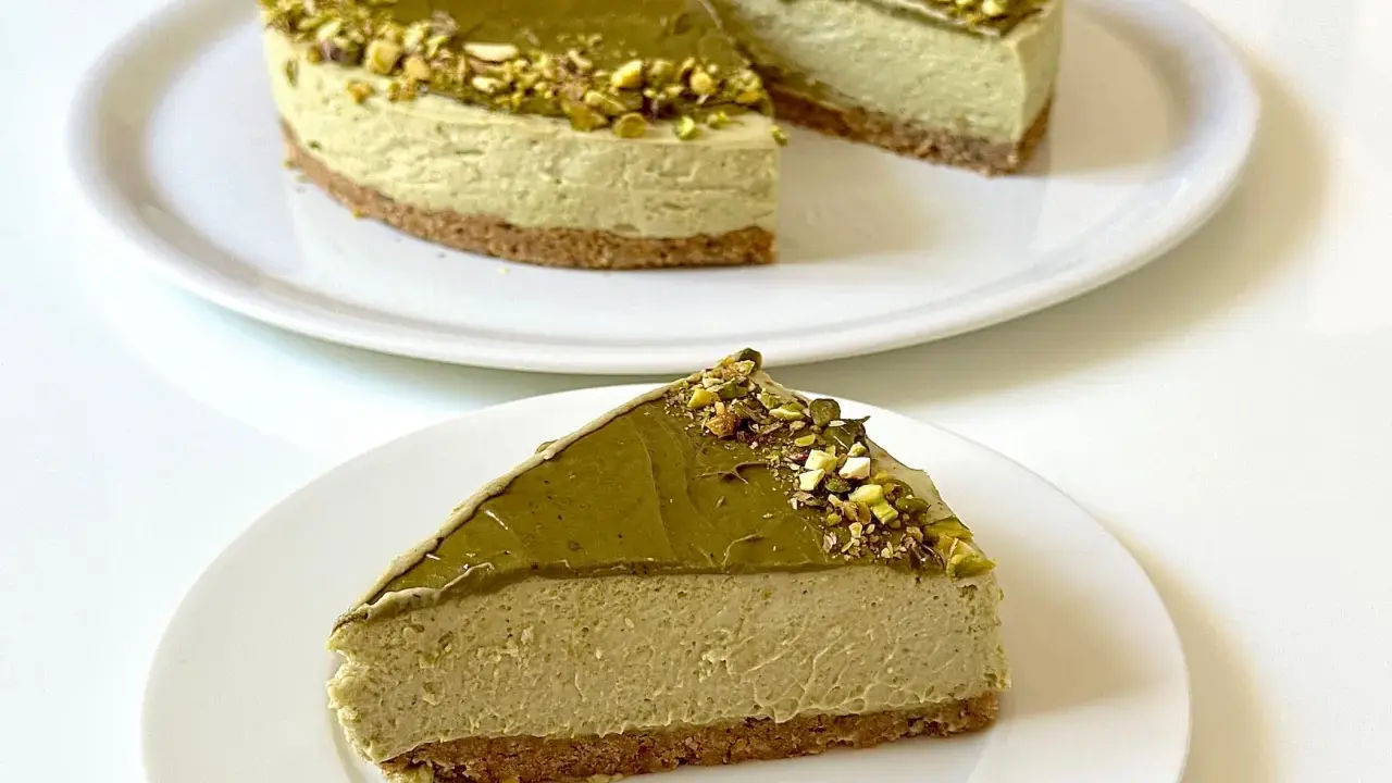 Una fetta di goloso cheesecake al pistacchio, con topping e granella di pistacchi, su un piatto bianco.