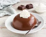 Budino al cioccolato liscio e lucido, guarnito con panna montata, su un piatto bianco. Accanto, due quadratini di cioccolato e una ciotolina di panna.