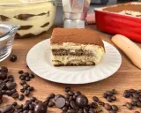 Una fetta di tiramisù con uova pastorizzate, cosparso di cacao, su un piatto bianco, circondato da chicchi di caffè e cioccolatini.