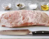 Un pezzo di girello di vitello crudo, pronto per essere cucinato, con coltello e ciotoline di condimenti sullo sfondo.