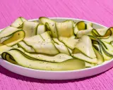 Fettine sottili di zucchine marinate crude, condite con pepe nero, disposte a spirale in un piatto bianco su un tavolo rosa acceso.