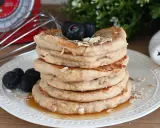 Pila di pancake con farina di avena, guarniti con mirtilli e fiocchi d'avena, su un piatto bianco con sciroppo d'acero.