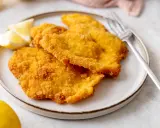 Due cotolette di pollo dorate e croccanti, servite su un piatto con spicchi di limone e una forchetta.