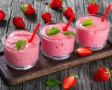 Tre bicchieri di spumone alle fragole bimby, guarniti con menta e fragole fresche, su un vassoio di legno.