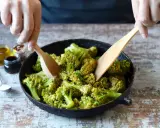 Mani che mescolano broccoli in padella con due palette di legno. Sullo sfondo, piccoli barattoli con olio, aceto balsamico e spezie.