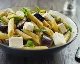 Piatto di pasta con melanzane in bianco, cubetti di formaggio fresco e foglie verdi, servito in una ciotola scura.