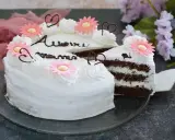 Una torta festa della mamma con glassa bianca, fiori rosa e cuori di cioccolato. La fetta tagliata rivela strati di torta al cioccolato e crema.
