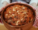 Un invitante pasta al forno appena sfornata, con crosta dorata e ripieno filante, tenuta con un canovaccio a quadretti rossi.