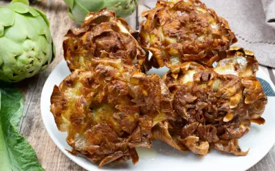 Carciofi alla giudia dorati e croccanti su un piatto bianco, con carciofi freschi sullo sfondo.