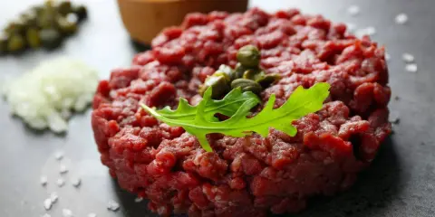 Tartare di manzo fresca, guarnita con capperi e rucola, pronta per essere gustata con tuorlo d'uovo e cipolla.