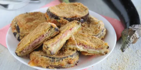 Cordon bleu di melanzane impanati e fritti, ripieni di prosciutto e formaggio filante, serviti su un piatto bianco.