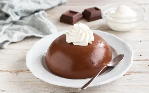 Budino al cioccolato liscio e lucido, guarnito con panna montata, su un piatto bianco. Accanto, due quadratini di cioccolato e una ciotolina di panna.