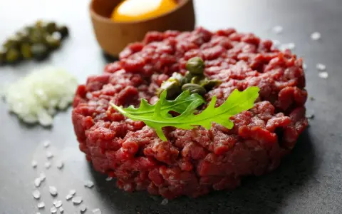Tartare di manzo fresca, guarnita con capperi e rucola, pronta per essere gustata con tuorlo d'uovo e cipolla.