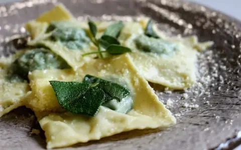Ravioli ricotta e spinaci, conditi con burro fuso e foglie di salvia fresca, spolverati di parmigiano grattugiato su un piatto elegante.