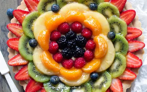 Torta alla frutta coloratissima, con fragole, kiwi, mandarini, lamponi e mirtilli disposti a raggiera su una base croccante.