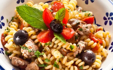 Pasta con pesce spada, pomodorini, olive nere e menta fresca in una ciotola decorata.