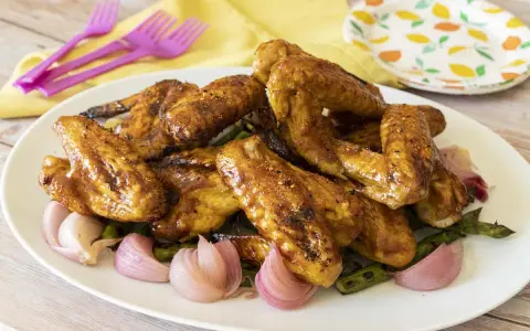Alette di pollo in padella, dorate e succulente, servite con cipolle rosse e asparagi su un piatto bianco.
