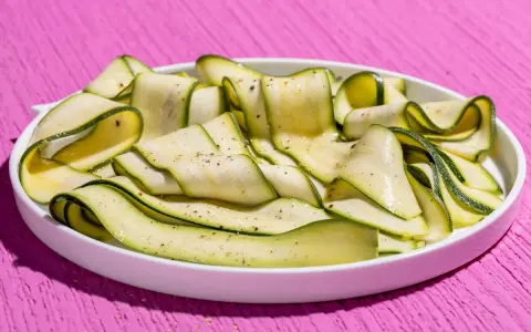 Fettine sottili di zucchine marinate crude, condite con pepe nero, disposte a spirale in un piatto bianco su un tavolo rosa acceso.