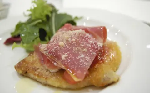 Cotoletta alla bolognese dorata, coperta da prosciutto crudo e parmigiano, con un contorno di insalata fresca.