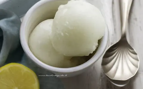 Due palline di sorbetto al limone senza albume in una ciotola bianca, con una fetta di limone e un cucchiaio decorato.