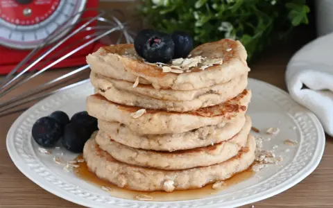 Pila di pancake con farina di avena, guarniti con mirtilli e fiocchi d'avena, su un piatto bianco con sciroppo d'acero.