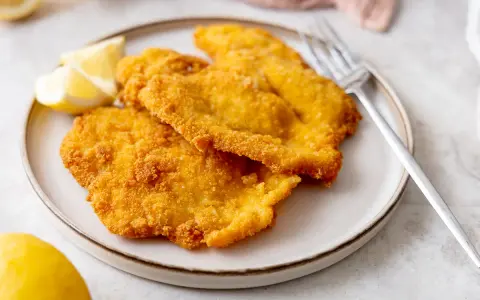 Due cotolette di pollo dorate e croccanti, servite su un piatto con spicchi di limone e una forchetta.