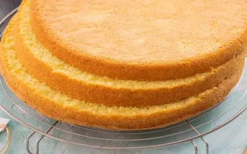 Tre soffici strati di pan di spagna senza glutine raffreddano su una griglia. Un'etichetta con scritto "senza glutine" è appesa alla griglia.