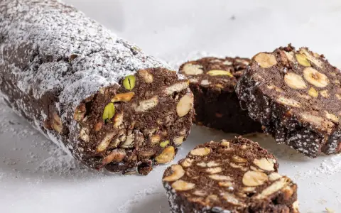 Salame di cioccolato senza uova, ricco di nocciole, mandorle e pistacchi, spolverato di zucchero a velo.