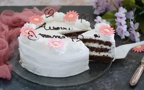 Una torta festa della mamma con glassa bianca, fiori rosa e cuori di cioccolato. La fetta tagliata rivela strati di torta al cioccolato e crema.