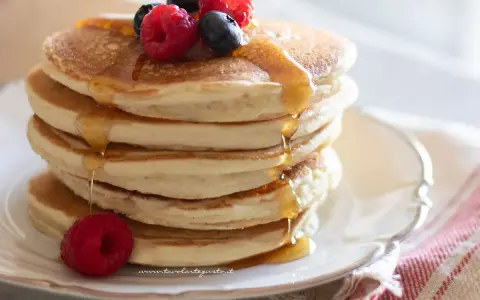 Pancakes senza glutine dorati, impilati e inzuppati di sciroppo, guarniti con lamponi e mirtilli freschi.