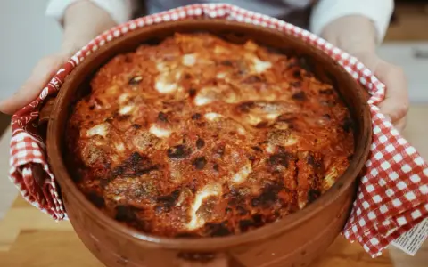 Un invitante pasta al forno appena sfornata, con crosta dorata e ripieno filante, tenuta con un canovaccio a quadretti rossi.
