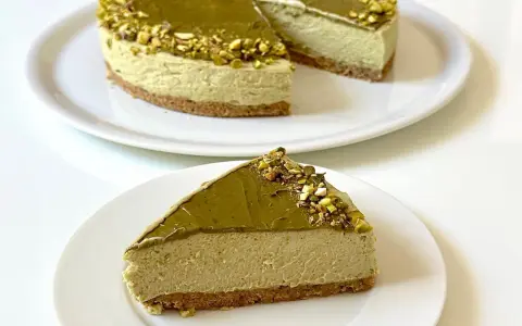 Una fetta di goloso cheesecake al pistacchio, con topping e granella di pistacchi, su un piatto bianco.