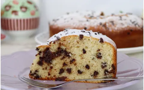 Una fetta di torta soffice con tante gocce di cioccolato, cosparsa di zucchero a velo, su un piatto lilla con una forchetta.