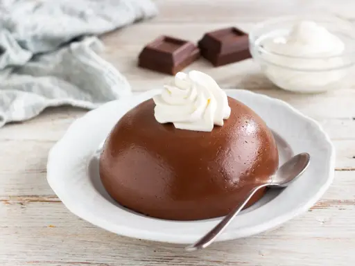 Budino al cioccolato liscio e lucido, guarnito con panna montata, su un piatto bianco. Accanto, due quadratini di cioccolato e una ciotolina di panna.